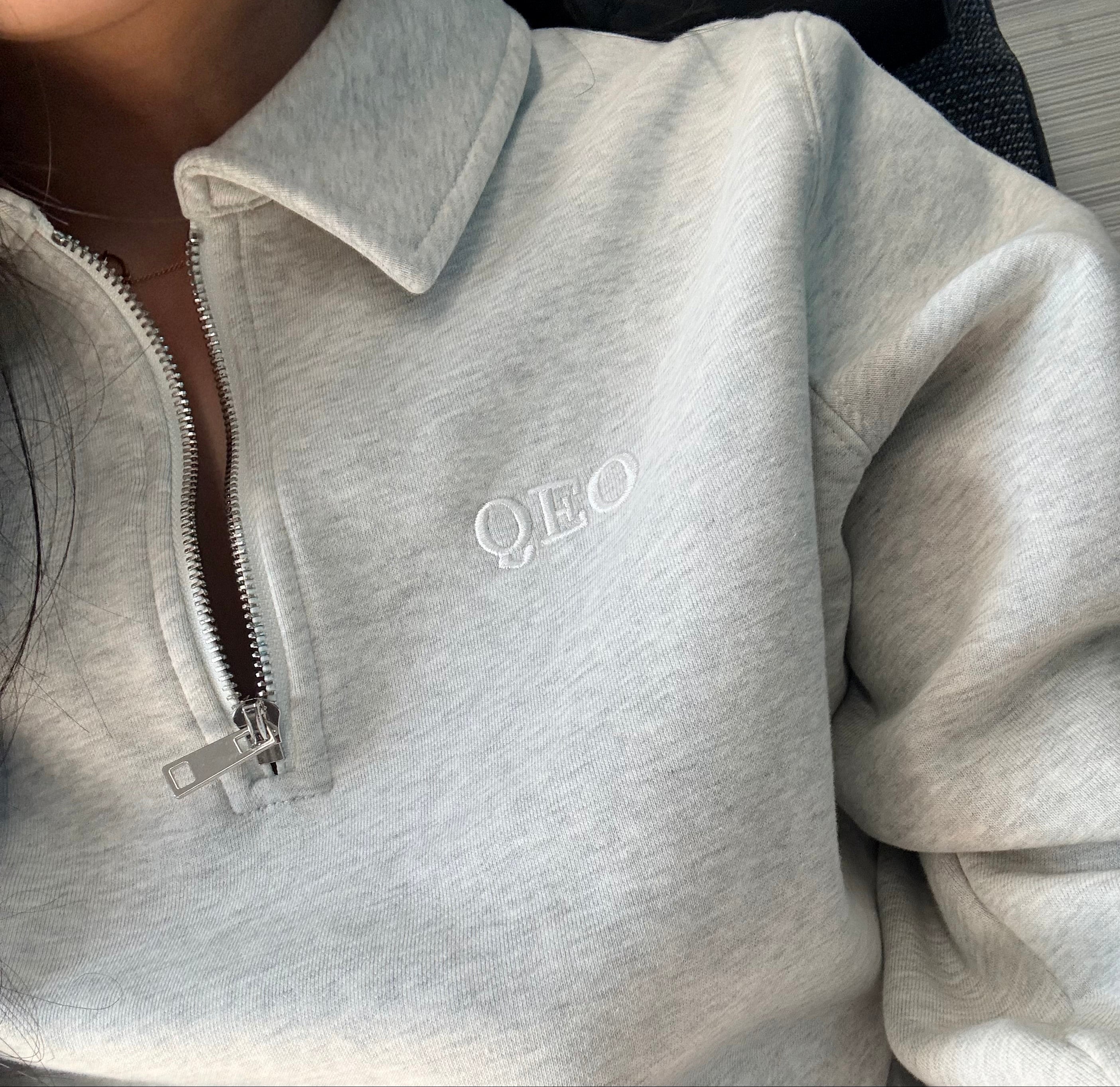 Grey quarter zip sweatshirt with 'QEO' logo on a neutral background