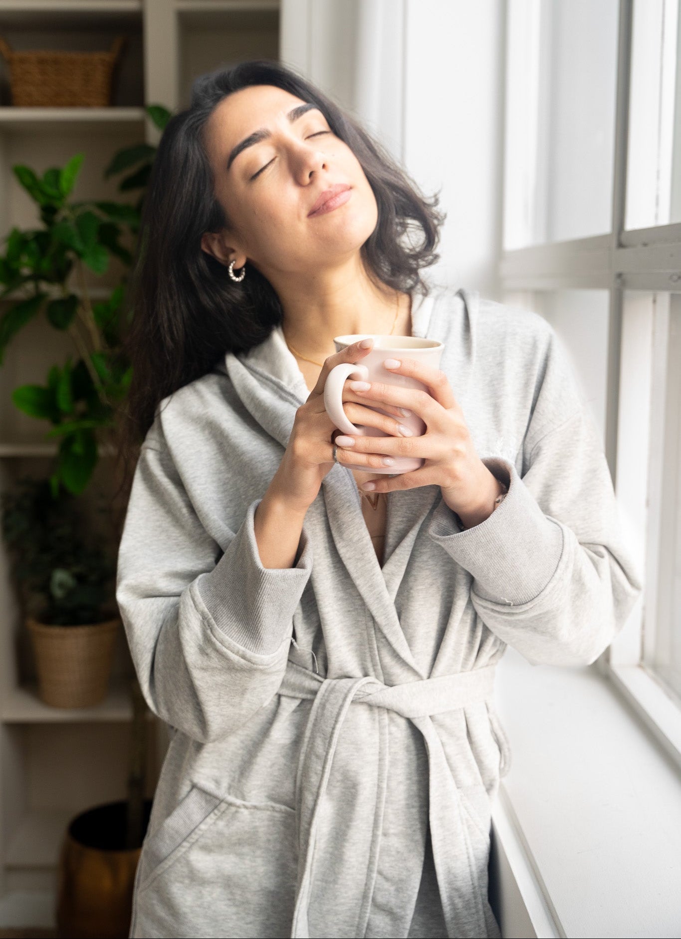 A woman wearing a light gray hooded robe stands by a window, holding a white mug with her eyes closed in a relaxed, cozy setting.